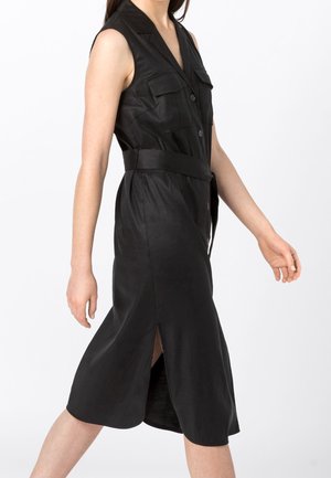 Vestido midi negro sin mangas con botones, bolsillos en el pecho con solapa, cinturón en la cintura y abertura lateral, llevado por una persona caminando sobre fondo blanco.