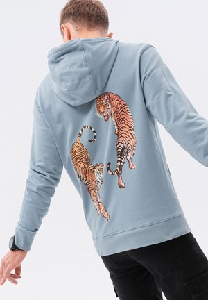 Persoon die een lichtblauwe hoodie draagt met twee oranje-zwarte tijgers op de rug gedrukt, staand tegen een effen witte achtergrond.