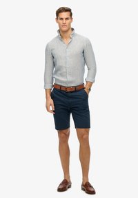 Camicia azzurro chiaro a maniche lunghe con righe verticali, pantaloni corti blu navy, cintura in pelle marrone e mocassini marrone scuro. Abbigliamento estivo casual.