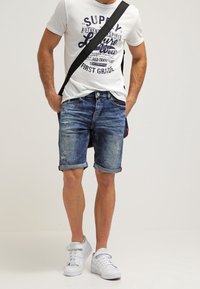 Shorts in denim blu con risvolti, caratterizzati da un look strappato. Abbinati a una t-shirt grafica bianca e sneakers bianche, con una borsa a tracolla nera.