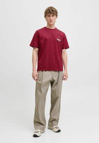 Bordeaux farvet bomulds t-shirt med korte ærmer og et enkelt print på venstre bryst, parret med beige plisserede bukser og cremede sneakers.