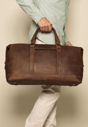 Borsa duffel in pelle marrone con doppi manici, tasca con zip, cuciture robuste e piedi metallici. Texture liscia e forma rettangolare.