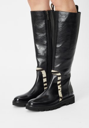 Bottes hautes en cuir noir avec des semelles épaisses et un détail en tissu rayé blanc près des chevilles, portées sur des jambes nues.