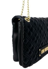 Czarna torebka pikowana z złotym łańcuszkiem, posiadająca teksturowaną skórzaną powierzchnię oraz prostokątny kształt. Widoczny detal z złotą tabliczką z logo.