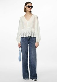 Blusa bianca con scollo a V, maniche a sbuffo e bordo in pizzo, abbinata a jeans blu a vita alta e una borsa leggera blu. 