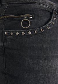 Svart denimtyg med en dragkedjeficka och en cirkulär metallackent, prydd med en rad silvernitar längs fickans kant.