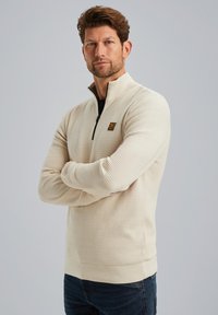 Beige ritssluiting trui met een gestructureerd stoffenpatroon, geribde manchetten en zoom. Bevat een klein logo op de borst. Draag het met donkere spijkerbroek.