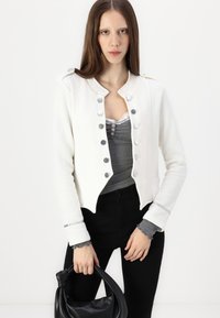 White cropped jacket with silver buttons, gray layered top, black pants, and textured black handbag. Smooth material with subtle design details.