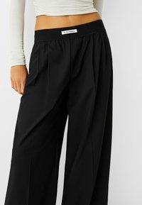 Mustat, korkeavyötäröiset housut, joissa on laskokset, joustava vyötärö ja etupuolella oleva etiketti, jossa lukee "KAIKENLAISIA KOKEMUKSIA".