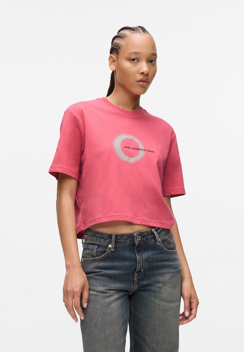 Ružičasti kratki majica od pamuka, s okruglim printom u sivoj boji i crnim tekstom "KARL LAGERFELD JEANS" centriranim na prednjem dijelu.