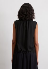 Persoon met krullend donker haar, die een mouwloos zwart topje met gehaalde zoom draagt, staand tegenover een eenvoudige witte muur.