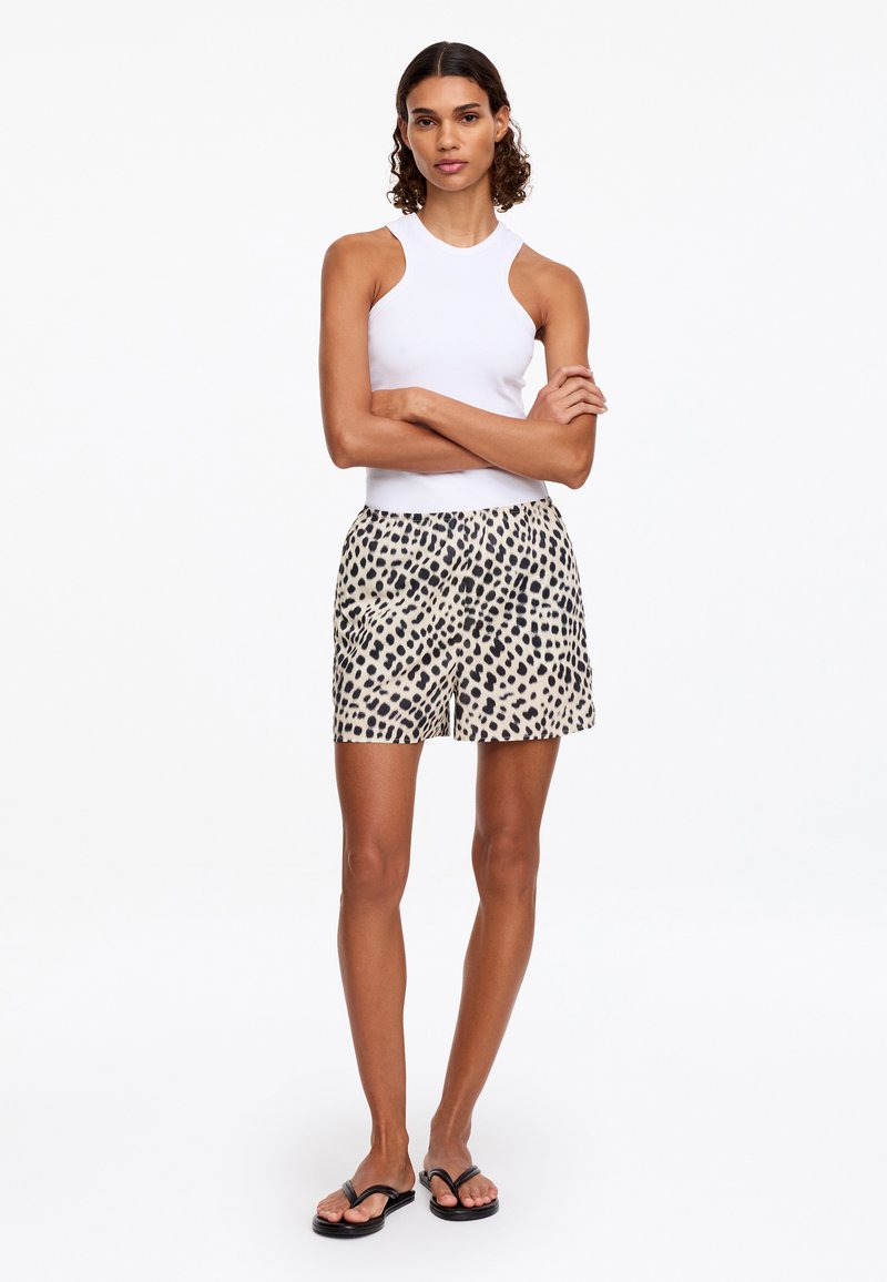 Femme debout, les bras croisés, portant un haut blanc sans manches, un short imprimé léopard et des tongs noires sur fond blanc.