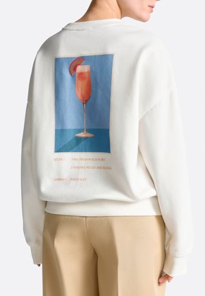 Person trägt weißen Pullover mit Grafik eines Pfirsichcocktailglases und Rezepttext auf der Rückseite, kombiniert mit beigen Hosen.