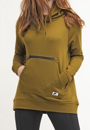 Vrouw draagt een olijfgroene hoodie met zak aan de voorkant en ritsdetail, handen in de zakken, gecombineerd met zwarte legging.