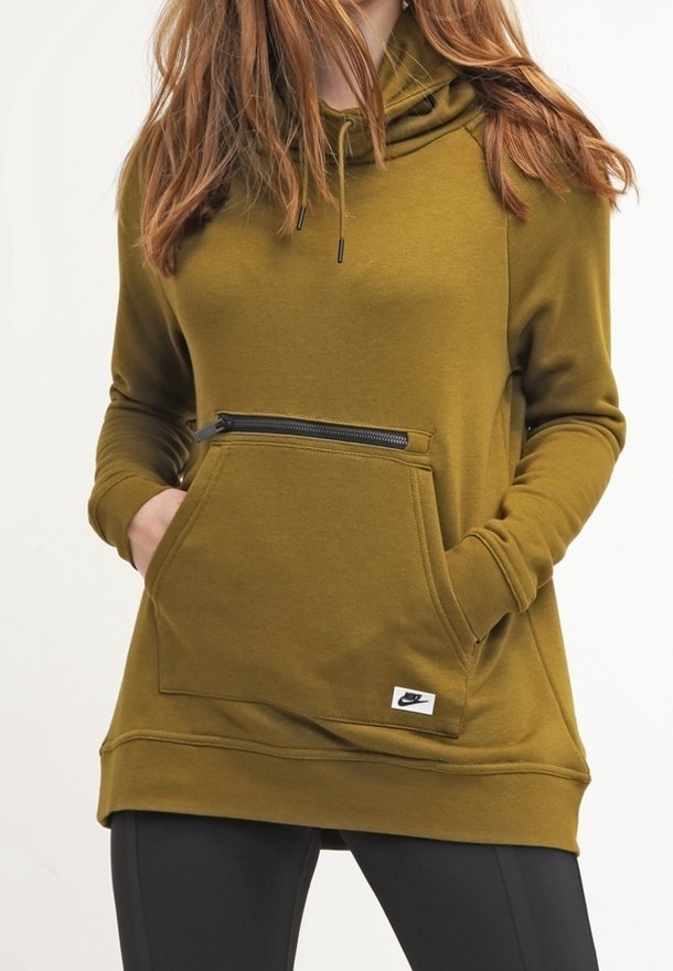Femme portant un sweat à capuche vert olive avec poche avant et détail zippé, mains dans les poches, associé à des leggings noirs.
