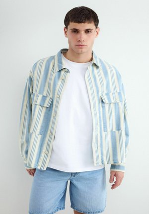 Jonge man draagt een wit T-shirt, een lichtblauw en crèmekleurig gestreept overhemd met knopen en lichtblauwe denim shorts, staand tegen een effen witte achtergrond.