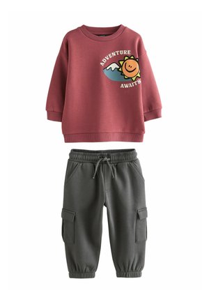 Felpa bordeaux con un design grafico di un sole e il testo "L'AVVENTURA TI ASPETTA"; abbinata a pantaloni cargo grigi con polsini elasticizzati e tasche.