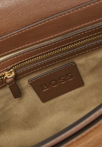 Indersiden af en brun lædertaske med en guldfarvet lynlås, glat ruskindtekstur og et prægget "BOSS"-logo på et matchende brunt mærkat.