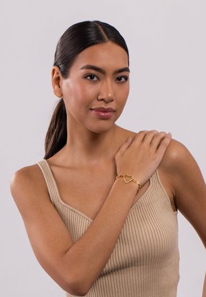 Goldarmband mit Herzdesign, ausgestattet mit einer Kettenverbindung. Das Model trägt ein beiges Ripptanktop und posiert mit einer Hand auf ihrer Schulter.