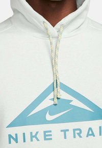 Hellblaues Hoodie mit einem dreieckigen Design, das ein großes Nike-Logo zeigt. Darunter steht in fetter Schrift "NIKE TRAIN". Texturierte Zugbänder in Blau und Gelb.