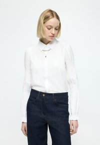 Bílá blůza s propracovaným límcem, dlouhými nafouklými rukávy s řasením, doplněná tmavými džínami z denimu s zlatým srdíčkovým detailem na pasu.