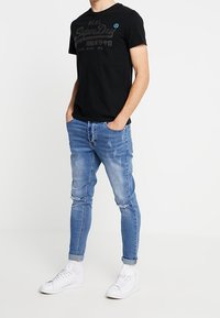Camiseta negra de manga corta con estampado gráfico, combinada con jeans ajustados desgastados de color azul claro y zapatillas blancas. Atuendo casual.