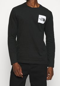 Svart långärmad t-shirt i bomull med en vit fyrkantig ficka med "The North Face"-logotyp. Mjuk textur, rund halsringning, smal passform.