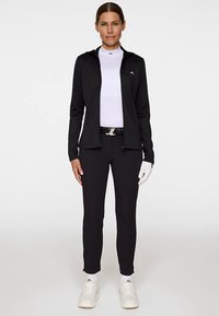 Svart zip-up jacka med en vit polokrage under, svarta byxor, vita skor och ett svart bälte med en silverdetalj. Vit handske på ena handen.