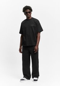 Schwarzes oversized T-Shirt mit kurzen Ärmeln, das einen dezenten Logo-Druck aufweist. Kombiniert mit lockeren schwarzen Hosen und Sneakers mit weißen Akzenten.