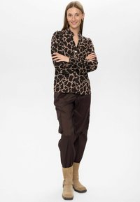 Donna in piedi con le braccia incrociate, che indossa una blusa con stampa animale marrone e nera, pantaloni cargo scuri e stivaletti beige su uno sfondo bianco.