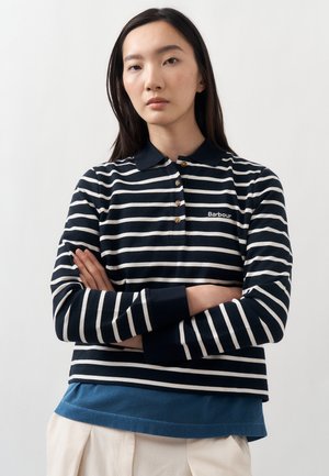 Młoda kobieta o długich ciemnych włosach nosi granatowo-białą, paskowaną koszulkę polo Barbour z kołnierzykiem, stoi ze skrzyżowanymi ramionami na jednolitym tle.