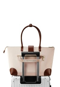 Beige tote taske med brune læderdetaljer, lynlåslukning og dobbelte håndtag, monteret på et kuffertgreb. Branding synlig på stroppen.