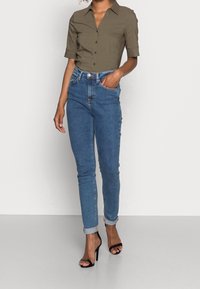Olijfgroene kortom mouwshirt met knoopsluiting, gecombineerd met hoge blauwe jeans. Zwart gestrapschoenen met hak maken de outfit compleet.