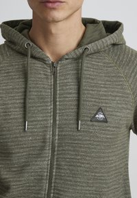 Sweat à capuche zippé vert olive en tissu texturé avec des rayures verticales, comportant un patch logo triangulaire noir et une capuche à cordons.