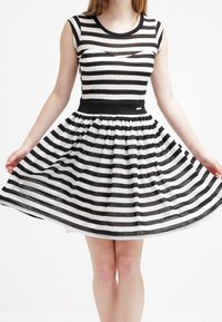 Robe à rayures noires et blanches avec des manches courtes, un corsage ajusté et une jupe évasée. Couche supérieure transparente et détails de ceinture noire.