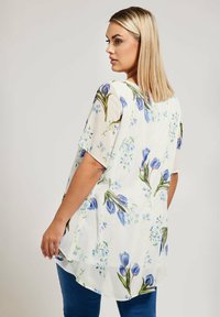 Blusa leggera e fluida color crema con motivi floreali blu. Presenta maniche corte, una vestibilità ampia e un orlo posteriore più lungo per una maggiore copertura.