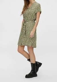 Grünes Blumenkleid mit kurzen Ärmeln, in der Taille gebunden und mit knielangem Saum. Kombiniert mit schwarzen Stiefeletten mit klobigen Sohlen.