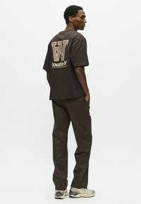 Hombre con una camiseta gráfica marrón oscuro, pantalones a juego y zapatillas beige claras, de pie de lado con las manos en los bolsillos sobre un fondo blanco.
