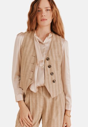 Gilet à rayures beiges avec cinq boutons, associé à une blouse en soie légère et froncée. Les accessoires comprennent un délicat collier en or.