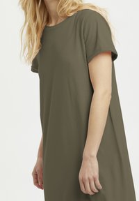 Olivgrünes T-Shirt-Kleid mit kurzen Ärmeln aus weichem Stoff, mit rundem Ausschnitt und lässiger Silhouette, das einen Teil des Arms zeigt.