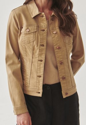 Veste en denim beige avec boutons en cuivre portée sur un haut beige assorti, associée à un pantalon noir. Modèle présenté du cou aux hanches.