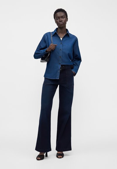 Femme portant une chemise en denim bleu foncé et un jean à jambes larges, tenant un sac à main noir avec une bandoulière chaîne, debout devant un fond uni.