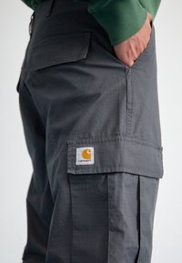 Harmaat cargo-housut, joissa on teksturoitu pinta, sivutasku ja Carhartt-logo oranssilla ja valkoisella tikkauksella.