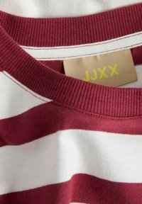 Close-up van een t-shirt met maroon en witte horizontale strepen, met een zichtbaar label waarop "JJXX" in het geel op een beige achtergrond staat.