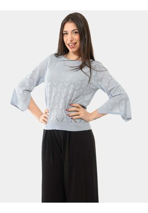 Donna con lunghi capelli castani che sorride, mani sui fianchi, indossa un maglione azzurro chiaro con motivo e pantaloni neri larghi su uno sfondo semplice.