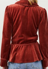 Chaqueta de terciopelo rojo óxido con cintura de peplum, cuello estructurado y mangas abullonadas, que presenta detalles cosidos a lo largo de la espalda y las mangas.