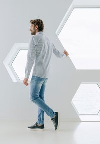 Pruhovaná košeľa s dlhým rukávom v svetlo modrej a bielej farbe, skombinovaná so svetlo modrými džínsami a tmavými teniskami. Hexagonálne okná na bielych stenách.