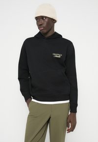 Άνδρας που φοράει μπεζ πλεκτό σκουφί, μαύρο φούτερ με το κείμενο "Originals Studio", λευκό μπλουζάκι κάτω από το φούτερ και ελιάς πράσινο παντελόνι, μπροστά σε λευκό φόντο.