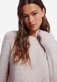 Maglione a maglia beige chiaro con polsini a coste e scollatura rotonda, con una consistenza morbida. Lunghi capelli castani ondulati cadono su una spalla.