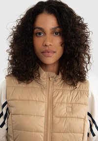 Béžová zateplená vesta s vysokým límcem, vertikálním prošíváním, předním zipem a malou nášivkou loga na levé straně. Měkký, texturovaný materiál.